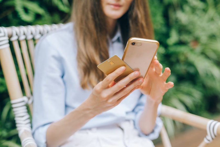 Mulher sentada ao ar livre segura um cartão bancário e o celular ao mesmo tempo.
